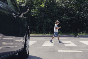 Low angle of car in front of pedestrian crossing and walking boy with smartphone